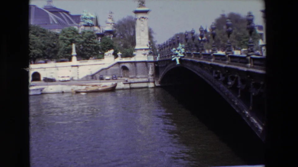 1969 Paris home-movie view of a low ornate bridge over the Seine, with moored boats along the quay and the Grand Palais roofline visible behind trees.