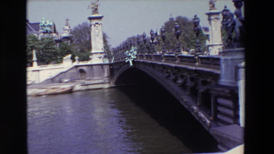 1969 Paris frame showing the Seine passing under an ornate low bridge, with stone bridge details, trees, and the Grand Palais roofline beyond the riverbank.