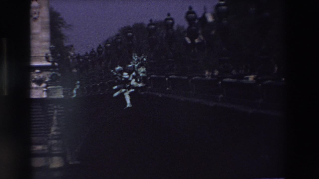 1969 Paris home-movie frame showing a pale stone statue or sculptural group illuminated against dark trees, lamppost silhouettes, and a deep twilight sky.