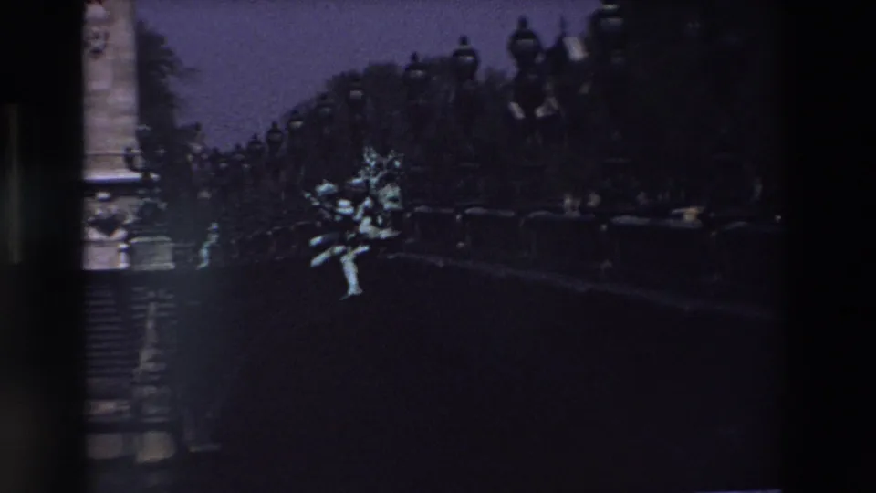 1969 Paris home-movie frame showing a pale stone statue or sculptural group illuminated against dark trees, lamppost silhouettes, and a deep twilight sky.