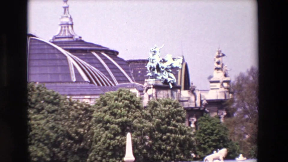 1969 Paris home-movie frame with green trees, distant ornate statuary, and a grand domed or glass-roofed building behind the monument area.
