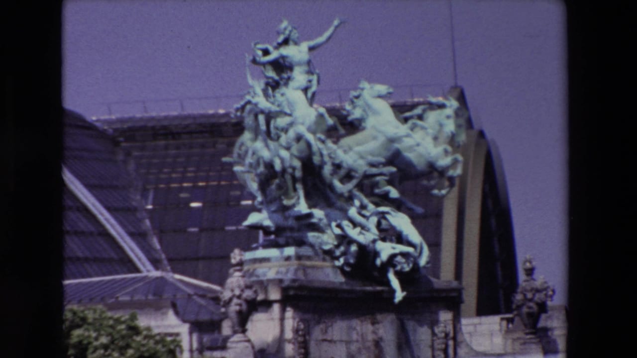 1969 Paris home-movie frame showing the L'Harmonie Triomphant bronze quadriga sculpture on a stone base with the Grand Palais roof behind it.