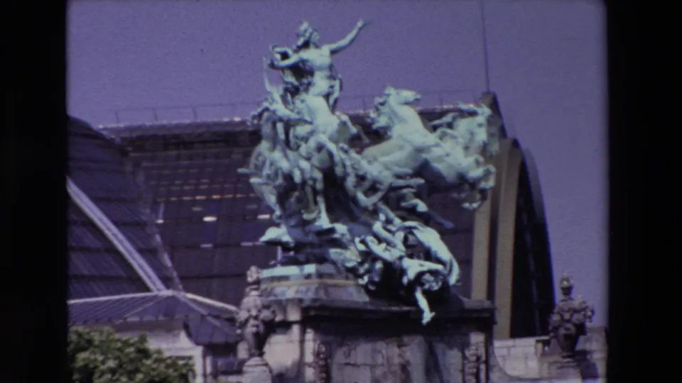 1969 Paris home-movie frame showing the L'Harmonie Triomphant bronze quadriga sculpture on a stone base with the Grand Palais roof behind it.