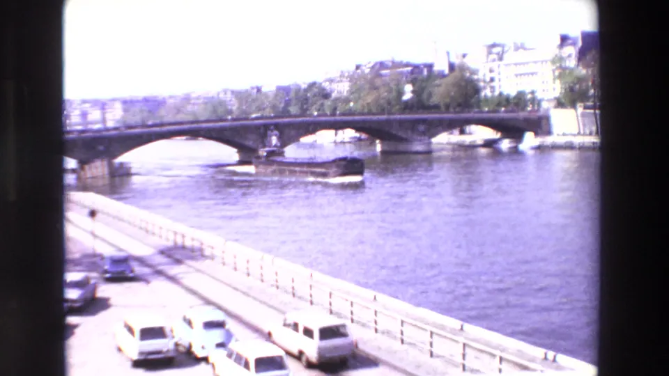 1969 Paris home-movie view across the Seine with a bridge, river traffic, embankment railings, parked cars, and riverside buildings in bright exposure.