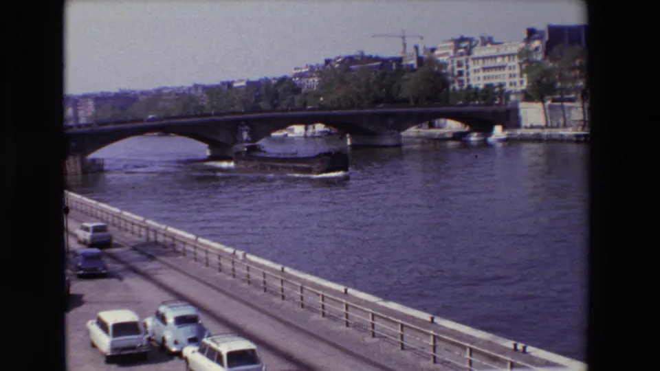 1969 Paris frame showing a wider Seine riverfront view with an arched bridge, a boat near the bridge, embankment railings, cars, and buildings along the far bank.