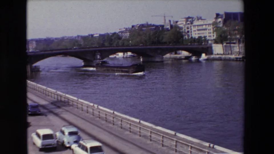 1969 Paris 8mm frame showing a large barge on the Seine near a stone arch bridge, with light-colored cars and a railing in the foreground.
