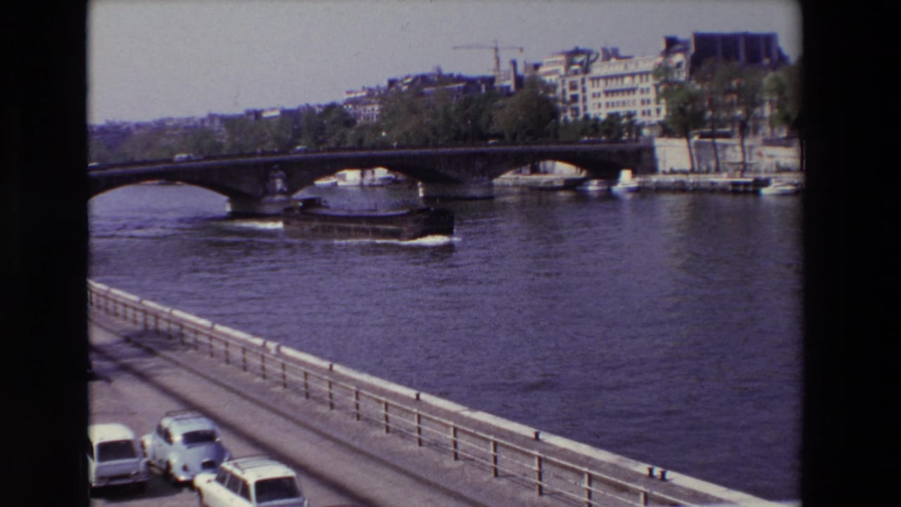 1969 Paris home-movie frame with a river barge below a stone bridge, embankment buildings and trees beyond, and parked or moving cars at the lower edge.