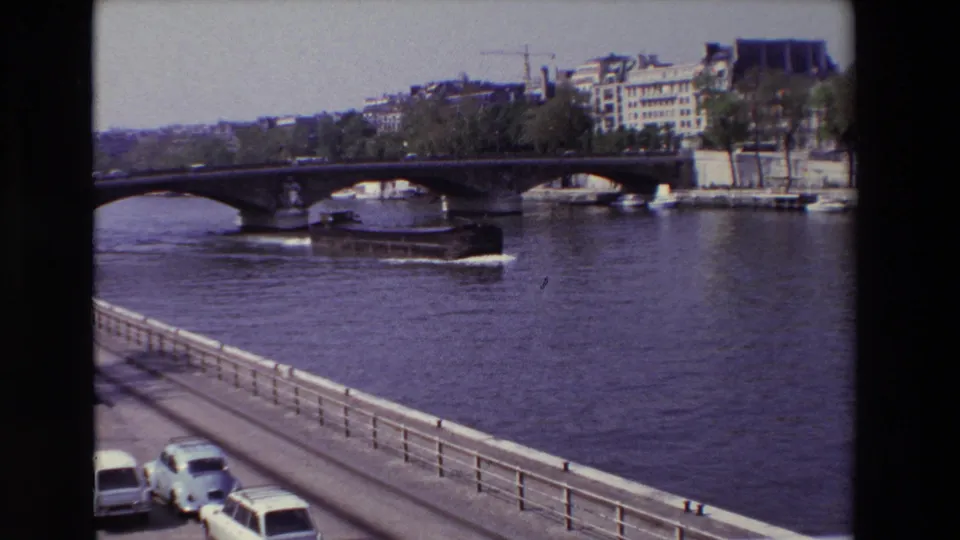 1969 Paris 8mm frame showing the barge farther along the Seine under the bridge, with foreground cars, railing, trees, buildings, and a crane visible in the distance.