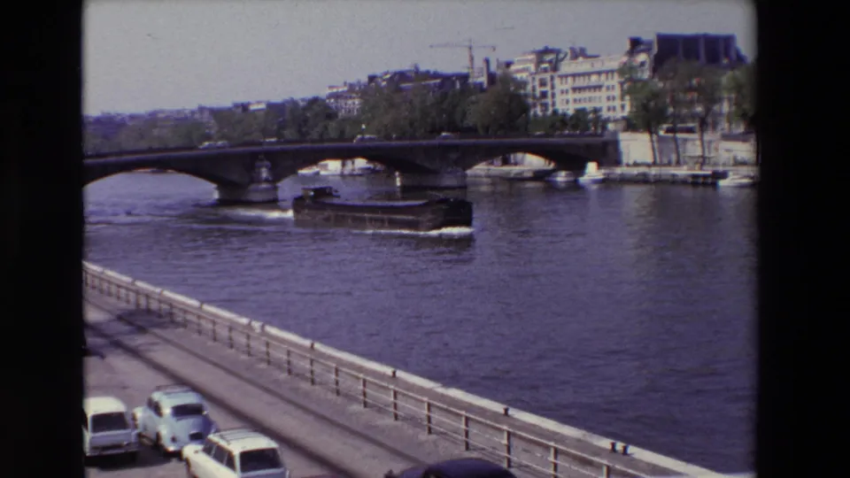 1969 Paris frame from a moving vehicle showing a barge on the Seine beyond a riverside railing, with a stone bridge, light cars, trees, and urban architecture in view.