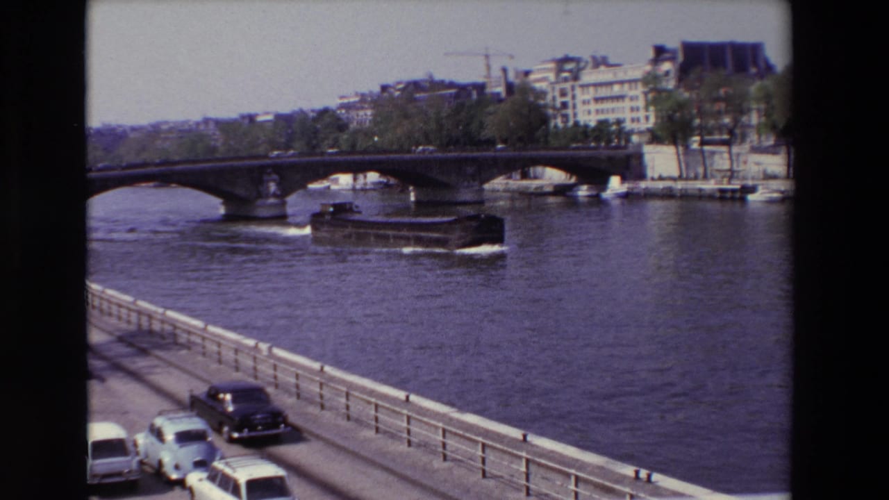 1969 Paris home-movie frame showing a boat on the Seine near an arched bridge, with parked and moving cars beside the river.