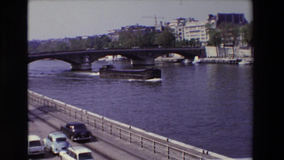 1969 Paris home-movie frame showing a boat on the Seine near an arched bridge, with parked and moving cars beside the river.