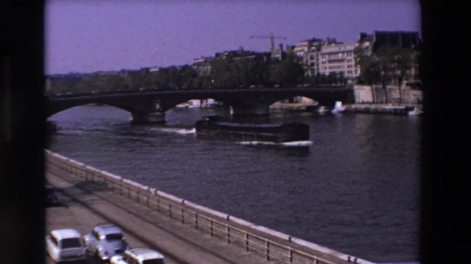 Faded 8mm frame from Paris in 1969 showing a Seine riverboat, arched bridge, riverbank buildings, and mid-century cars along the road.