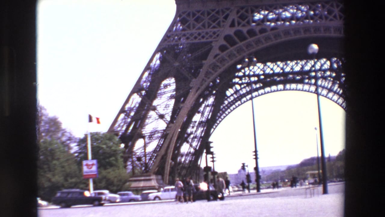 1969 Paris 8mm frame showing the Eiffel Tower from below, with pedestrians, period cars, traffic lights, a French flag, trees, and a distant horizon near the tower base.