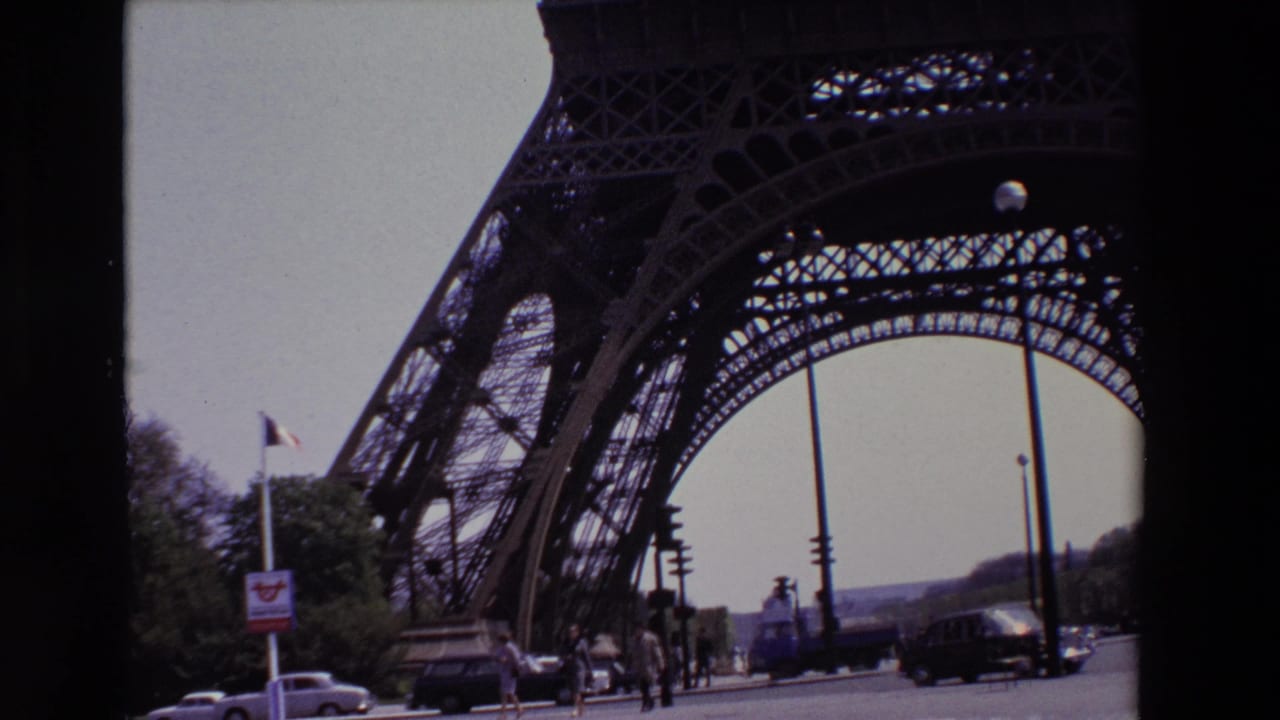 Eiffel Tower lower arch with pedestrians, cars, a flagpole, and street lights beneath an overcast sky.