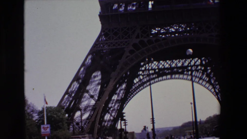 1969 Paris frame showing the Eiffel Tower's iron lattice and lower arch from a visitor perspective, with a French flag, trees, street signal, and lamps visible below.