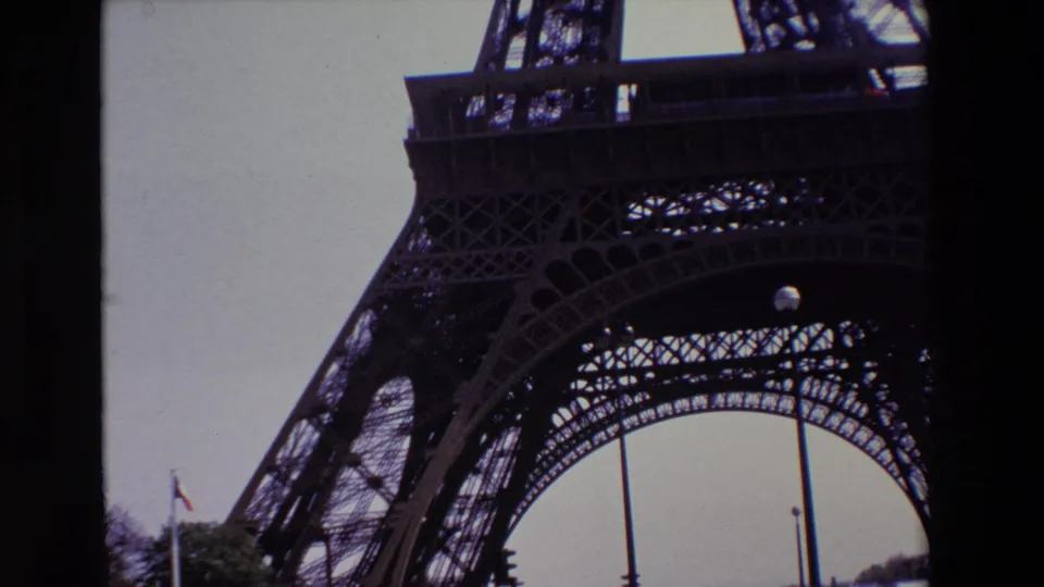 1969 Paris 8mm frame of the Eiffel Tower base and lower deck from below, with its iron lattice, French flag, trees, street fixtures, and muted cloudy sky.