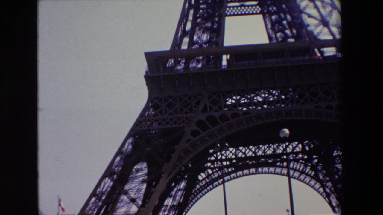 1969 Paris home-movie frame looking up at the Eiffel Tower's iron lattice and lower arch against a pale sky, with dark 8mm borders and a small French flag near the bottom left.
