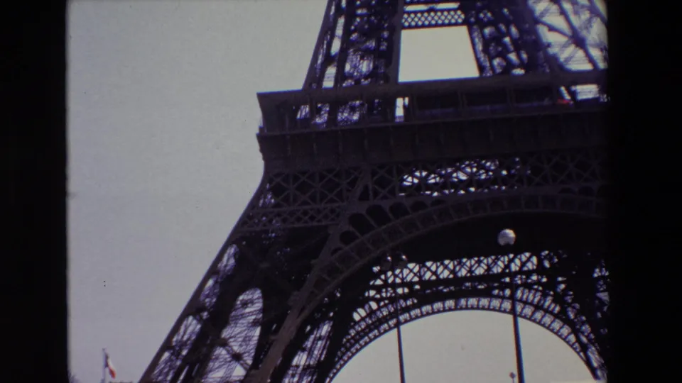 1969 Paris home-movie frame looking up at the Eiffel Tower's iron lattice and lower arch against a pale sky, with dark 8mm borders and a small French flag near the bottom left.