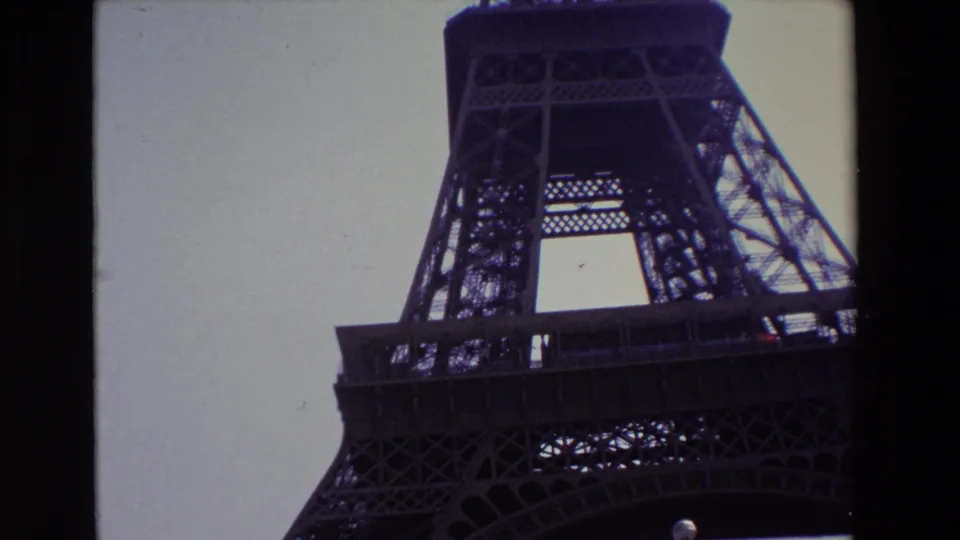 1969 Paris home-movie frame with the camera angled upward along the Eiffel Tower leg, showing the platform, latticework, lower arch, and dark film edge.