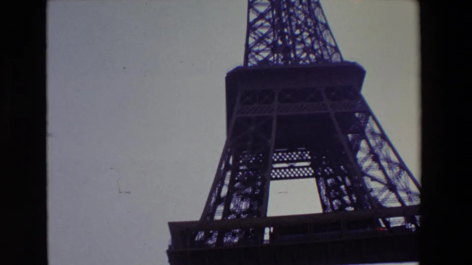 1969 Paris home-movie frame showing the Eiffel Tower from below, with the iron lattice and platform area silhouetted against a pale sky.