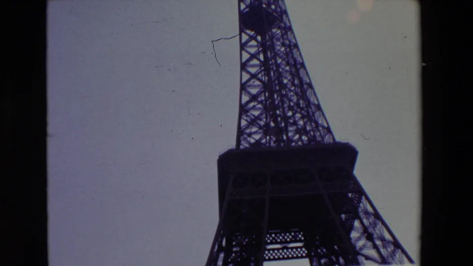 1969 Paris home-movie frame showing a closer low-angle view of the Eiffel Tower platform and lattice, with scratches and muted color visible in the scan.
