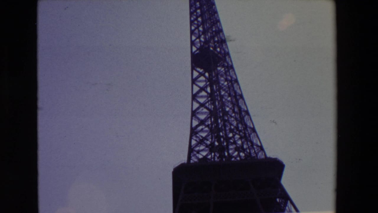 1969 Paris 8mm frame showing the lower Eiffel Tower in dark silhouette, cropped close against a muted sky with visible film-strip edges and grain.