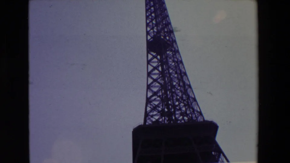 1969 Paris 8mm frame showing the lower Eiffel Tower in dark silhouette, cropped close against a muted sky with visible film-strip edges and grain.
