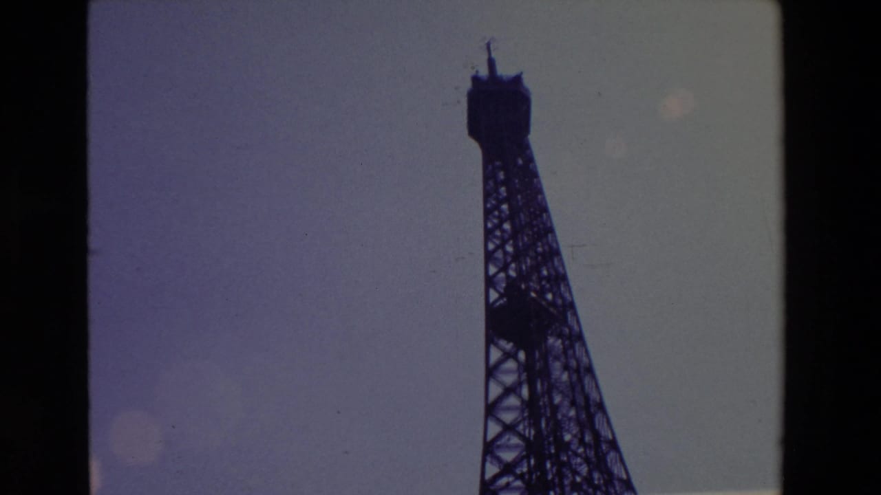 Upper Eiffel Tower silhouette with spire against a pale sky and visible film wear.