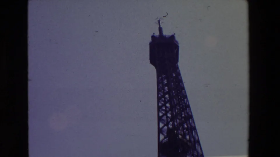 1969 Paris 8mm frame with the Eiffel Tower upper structure and spire silhouetted near the right edge, set against a faded sky with analog film texture.