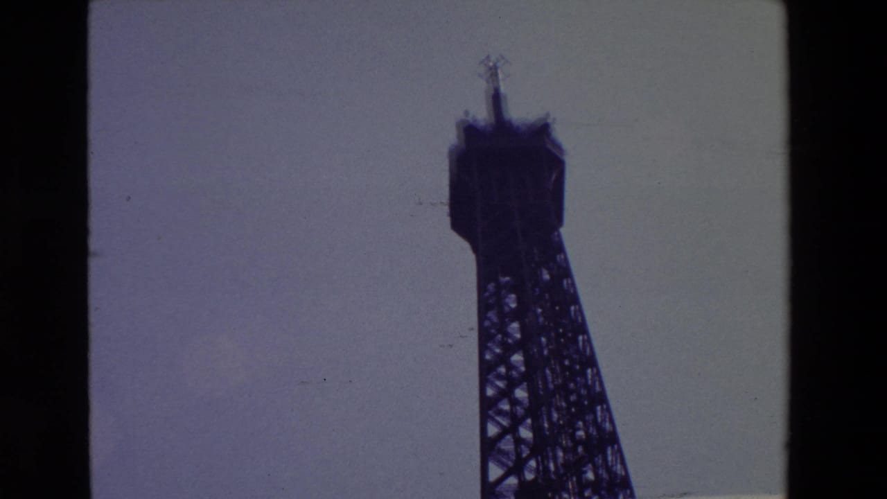 1969 Paris home-movie frame showing the Eiffel Tower's upper structure in dark silhouette against a pale sky, with black film borders visible.