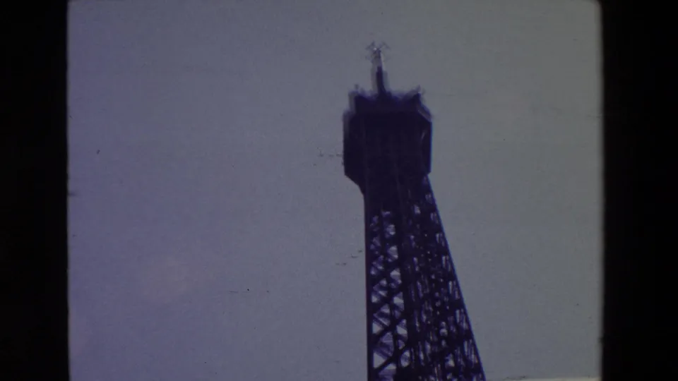 1969 Paris home-movie frame showing the Eiffel Tower's upper structure in dark silhouette against a pale sky, with black film borders visible.