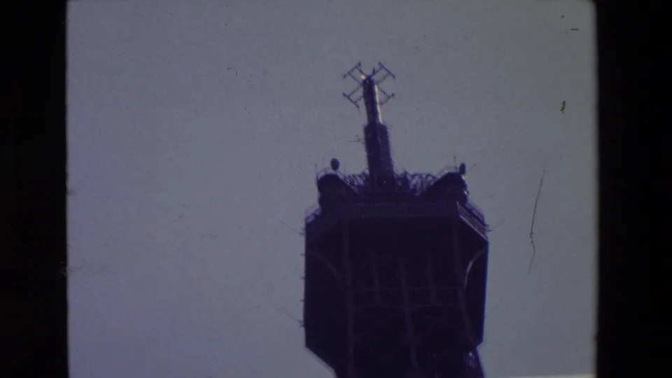 Closer 1969 Paris home-movie view of the Eiffel Tower's top platform and antenna elements, rendered in soft focus with purple-toned film color.