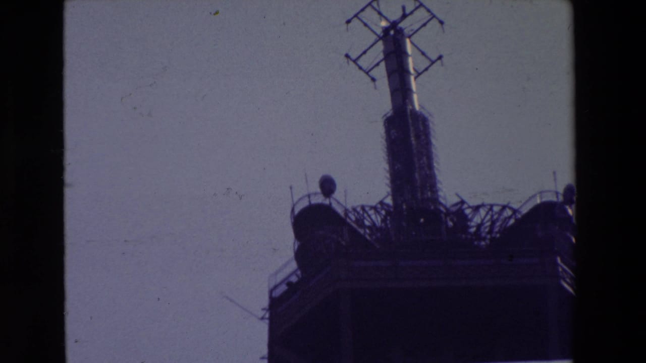 1969 Paris 8mm frame showing the Eiffel Tower's upper platform, central radio mast, and antenna details silhouetted against a pale sky.
