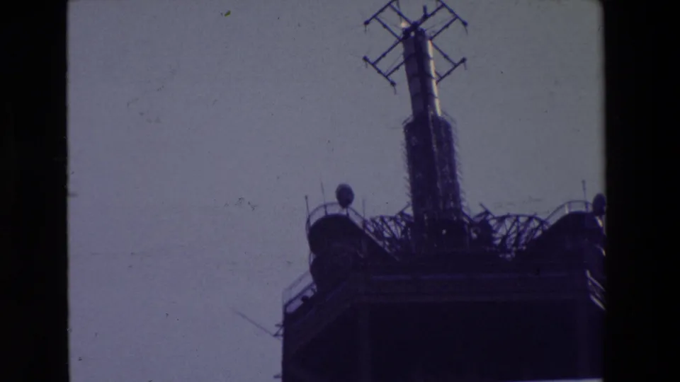 1969 Paris 8mm frame showing the Eiffel Tower's upper platform, central radio mast, and antenna details silhouetted against a pale sky.