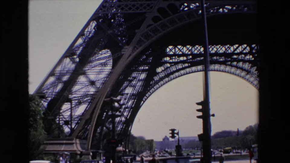 1969 Paris home-movie view under the Eiffel Tower, showing the curved iron arch, traffic lights, pedestrians, cars, and a tree-lined street beyond.