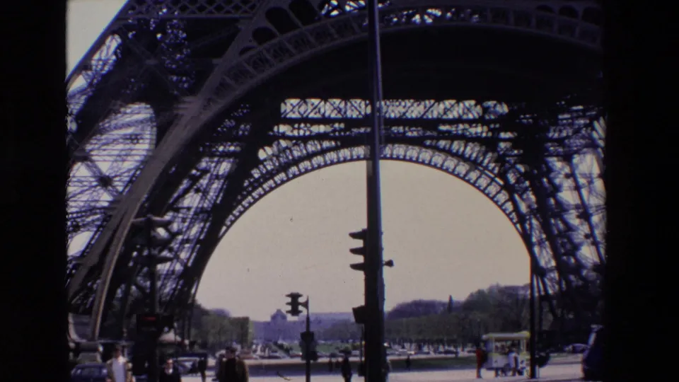 1969 Paris footage frames the Eiffel Tower arch above pedestrians, light-colored cars, traffic signals, trees, and distant urban buildings.