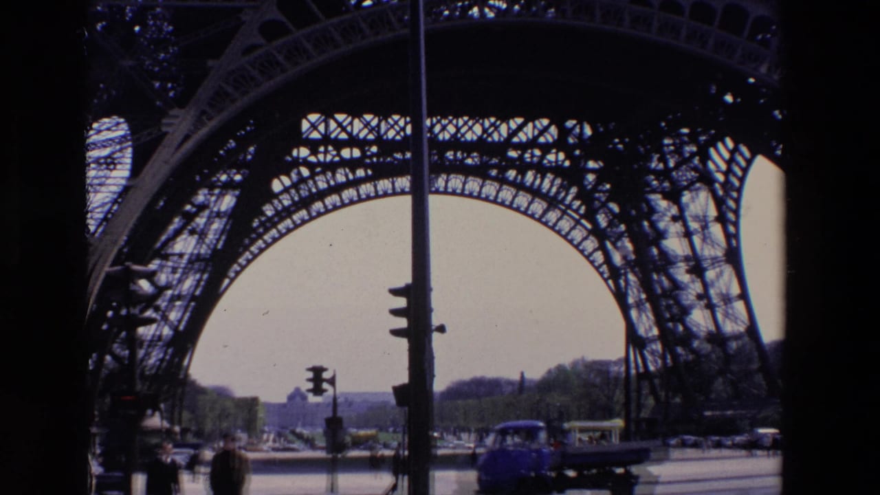 1969 Paris home-movie frame showing road traffic, pedestrians, and street signals seen through the iron lattice arches near the Eiffel Tower base.