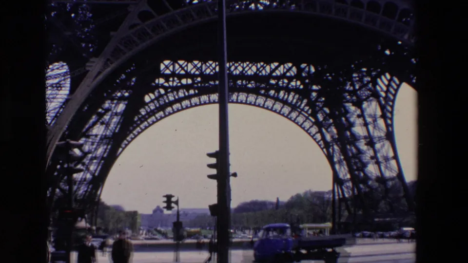 1969 Paris home-movie frame showing road traffic, pedestrians, and street signals seen through the iron lattice arches near the Eiffel Tower base.