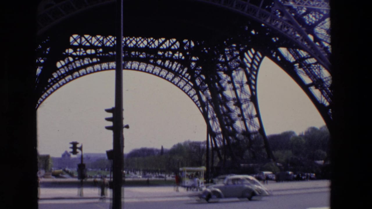 1969 Paris frame with the Eiffel Tower ironwork overhead, cars on the road, pedestrians near the sidewalk, and trees in the distance.