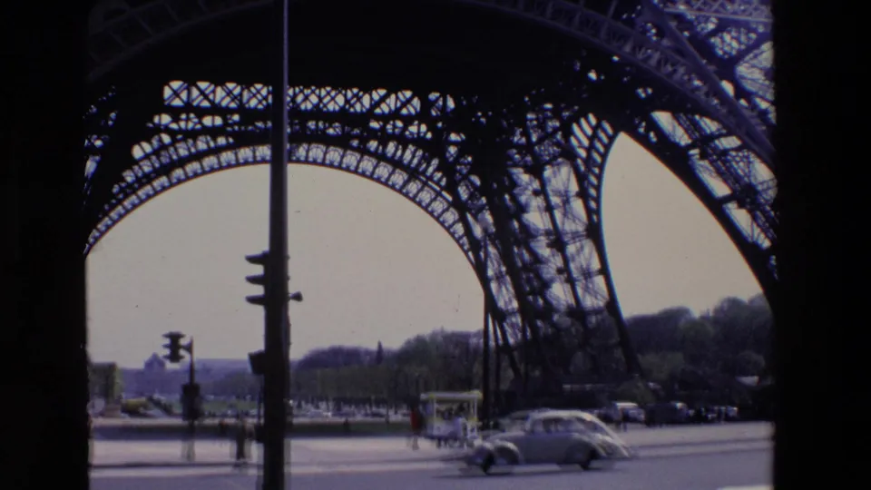 1969 Paris frame with the Eiffel Tower ironwork overhead, cars on the road, pedestrians near the sidewalk, and trees in the distance.