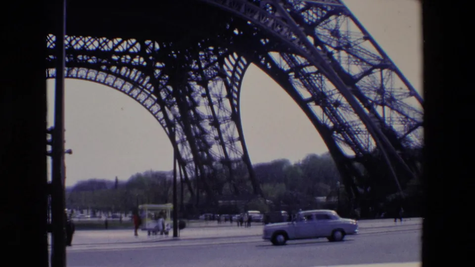 1969 Paris home-movie view of a light-colored car driving past the Eiffel Tower lattice with pedestrians, traffic signals, and roadside structures nearby.