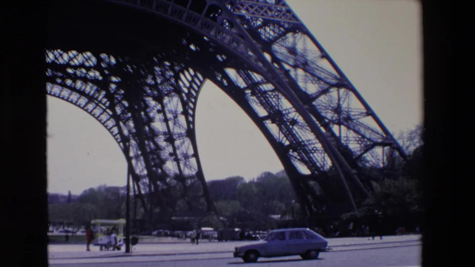 1969 Paris frame showing a car traveling along the road beneath the Eiffel Tower structure, with pedestrians, sidewalk activity, and trees in the background.