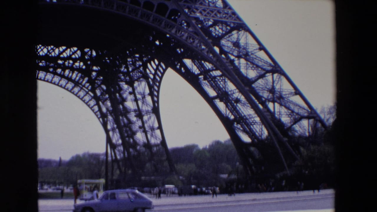 1969 Paris home-movie frame looking up at the Eiffel Tower iron lattice from below, with a small car, pedestrians, pavement, and distant trees at the base.