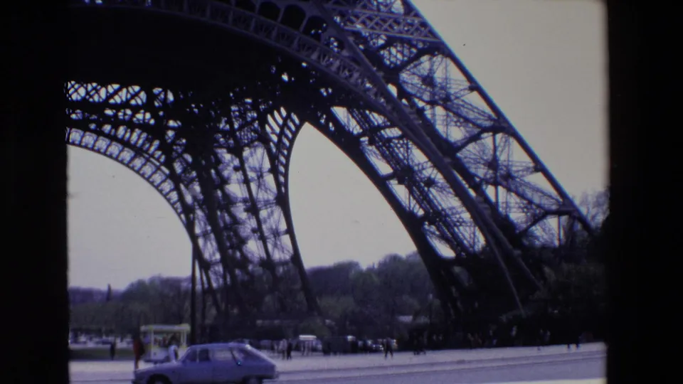 1969 Paris home-movie frame looking up at the Eiffel Tower iron lattice from below, with a small car, pedestrians, pavement, and distant trees at the base.