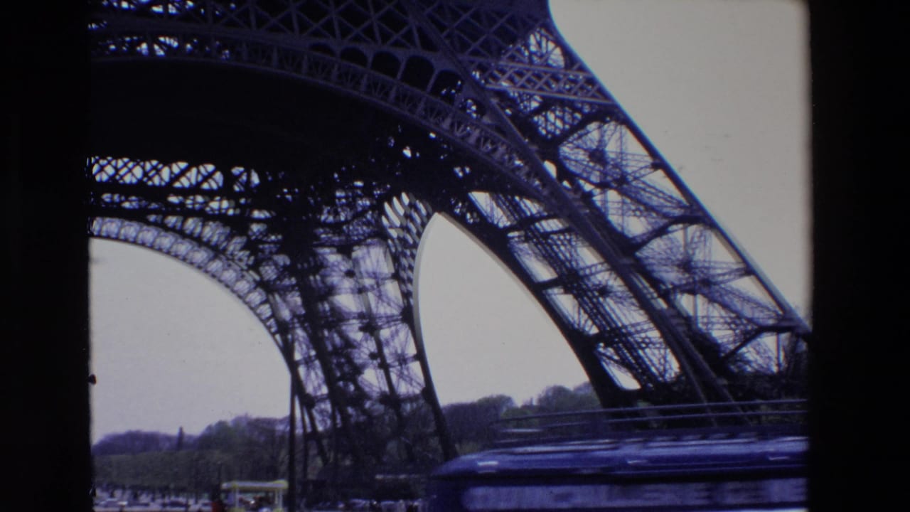 1969 Paris frame of the Eiffel Tower seen from beneath its iron structure, with a blurred vehicle passing across the lower foreground and small pedestrians near the base.