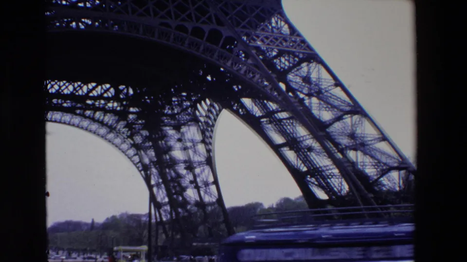 1969 Paris frame of the Eiffel Tower seen from beneath its iron structure, with a blurred vehicle passing across the lower foreground and small pedestrians near the base.