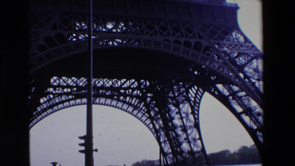 1969 Paris home-movie frame showing the Eiffel Tower ironwork from below, framed by a vertical pole, pale sky, and dark structural arches.