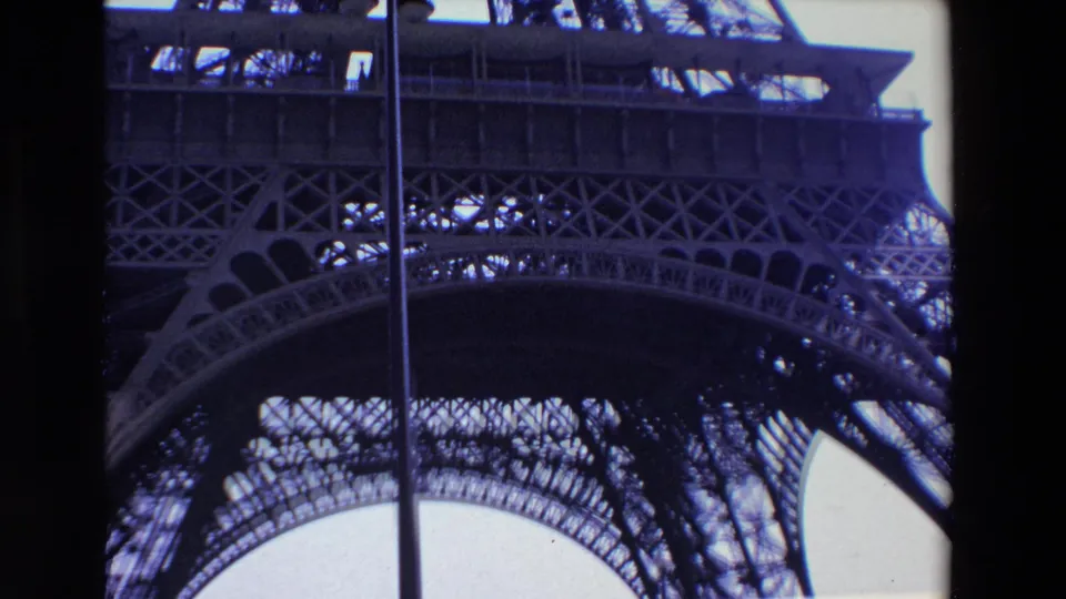 1969 Paris frame of the Eiffel Tower lower lattice and arching support seen from below, with a vertical pole in the foreground and pale sky behind the metalwork.