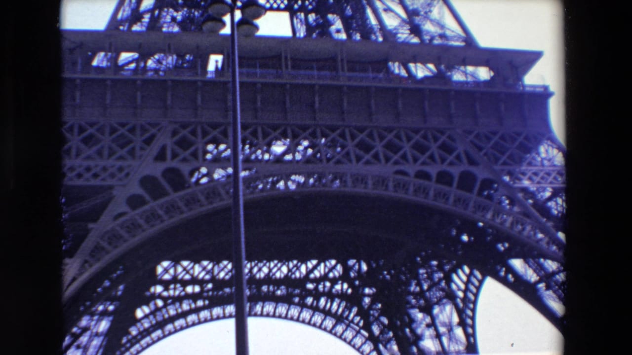 1969 Paris 8mm frame showing a low-angle view of the Eiffel Tower's lower lattice and arch structure against a bright sky, with a vertical lamppost in front.