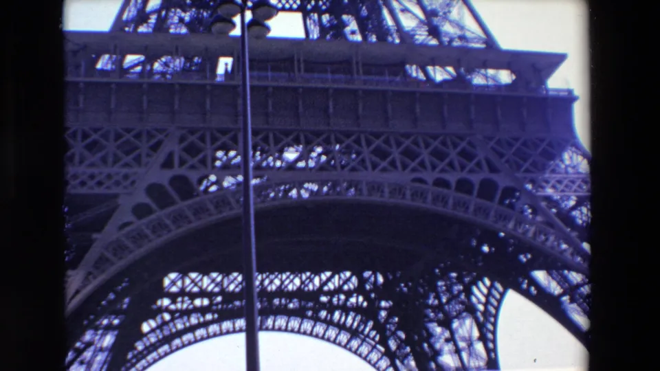 1969 Paris 8mm frame showing a low-angle view of the Eiffel Tower's lower lattice and arch structure against a bright sky, with a vertical lamppost in front.